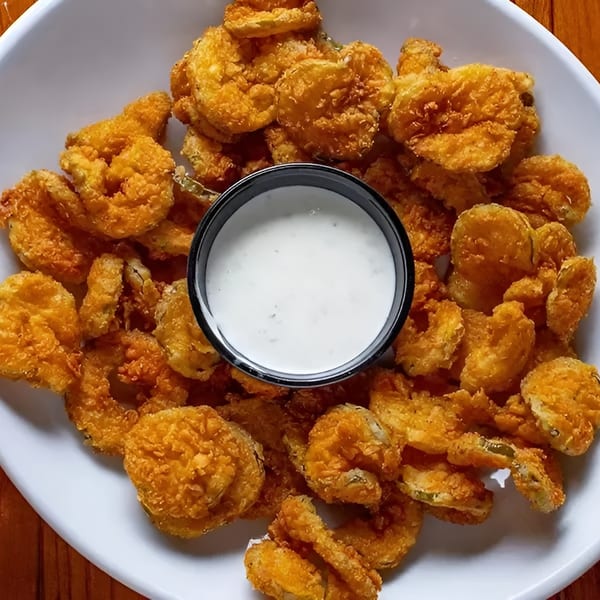 A white plate filled with golden-brown fried pickles and a small cup of ranch dressing.