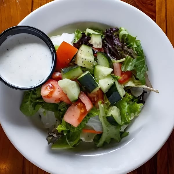 A fresh side salad with mixed greens, tomatoes, and cucumbers, served with a side of ranch dressing.